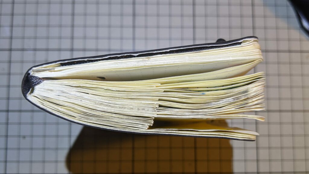 Side view of a well-used, thick notebook with cream-colored pages in a dark cover resting on a grey gridded cutting mat.