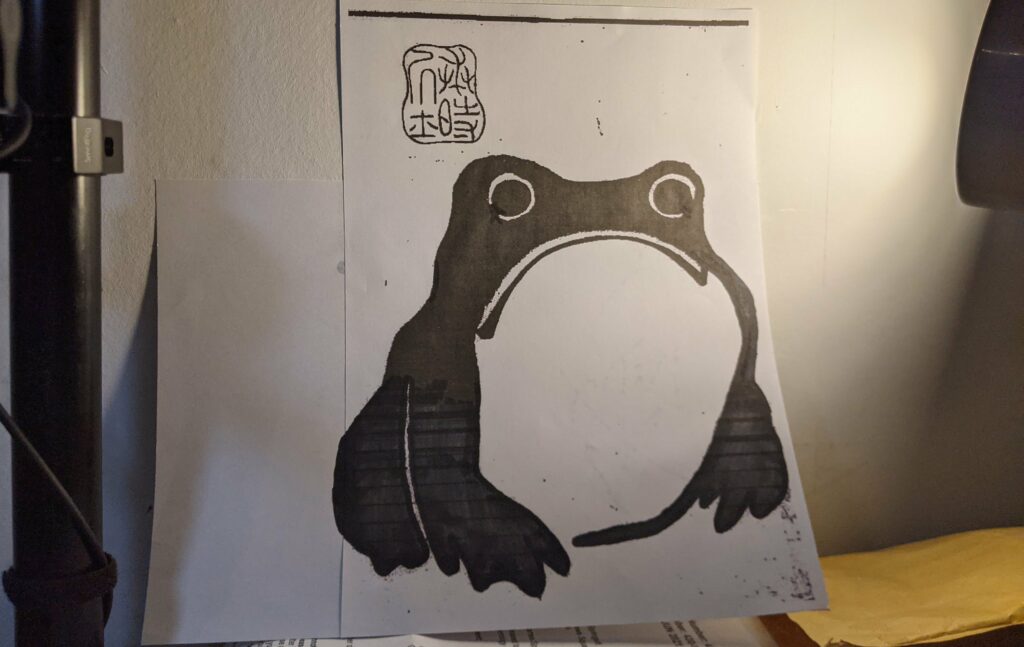 Photograph of sad frog drawing being held up in a room with red squiggle doodle visible at top