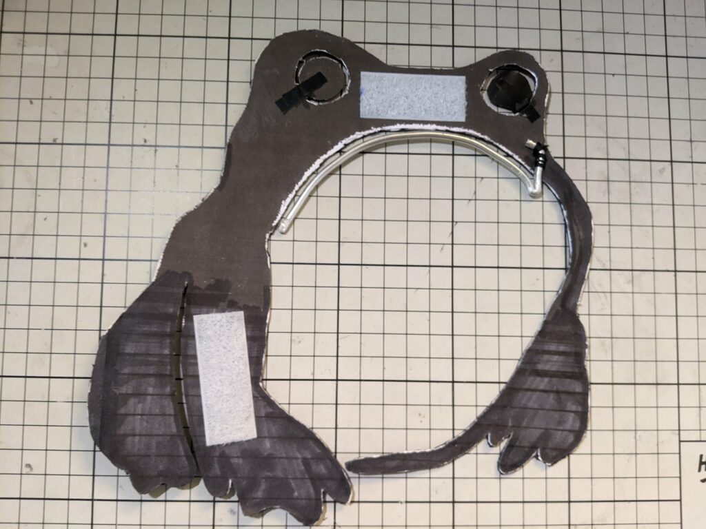 Dark puppet with frog-like features, white eye patches and curved mouth on tiled background