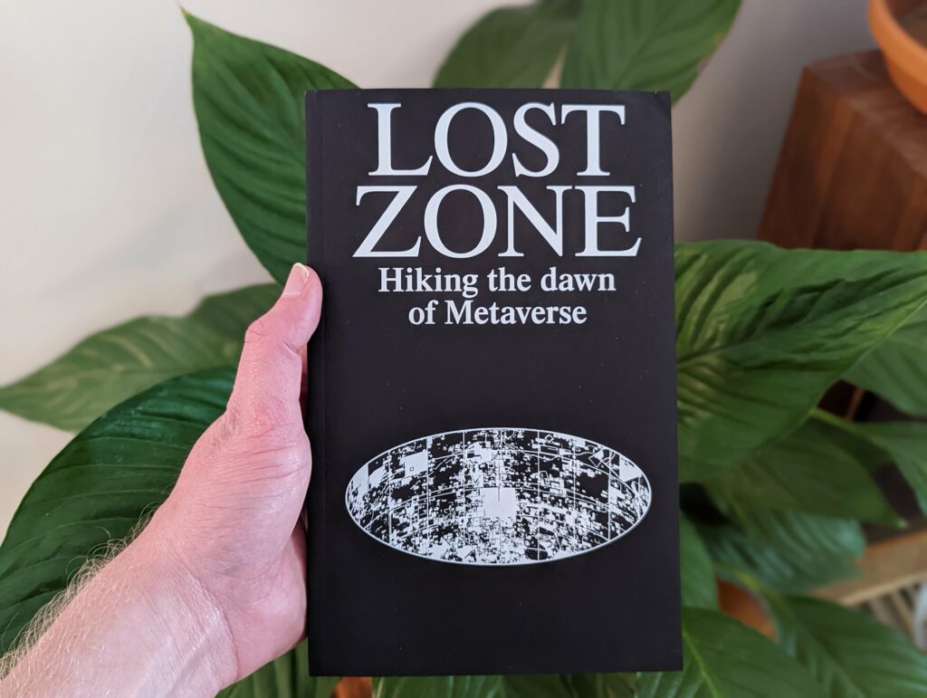 A hand holding a black book titled LOST ZONE: Hiking the dawn of Metaverse against a background of large green indoor plant leaves.