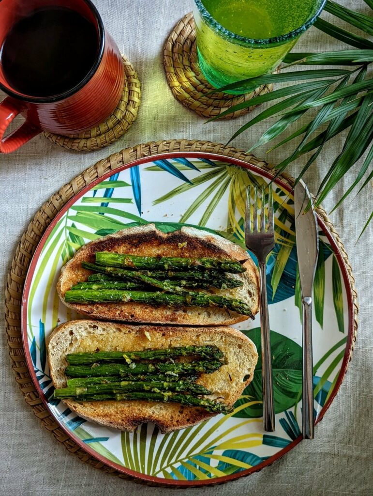 Grilled asparagus on sourdough toast on a tropical leaf plate, beside a red mug, green drink, and palm fronds.