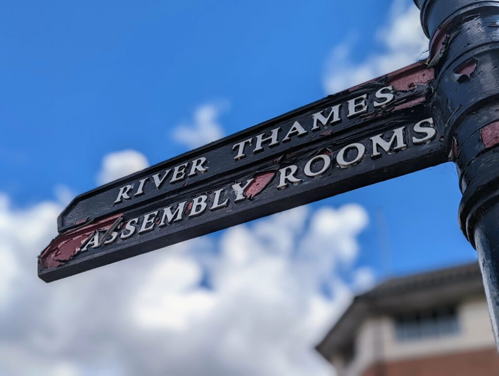A black fingerpost sign with peeling paint for the River Thames and Assembly Rooms against a blue sky with white clouds.