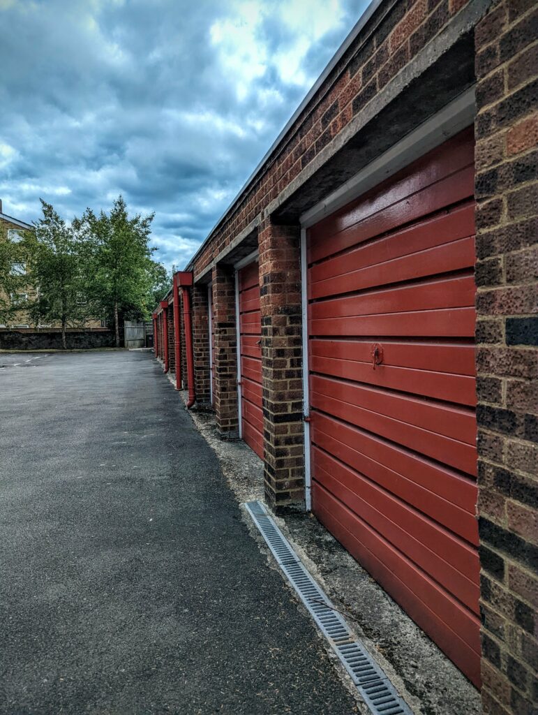 A row of red horizontal-slatted garage doors set into a brick building beside an asphalt lot under a cloudy sky.