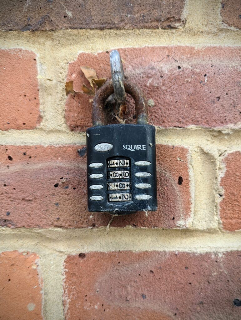 A weathered black Squire four-dial combination padlock with a rusty shackle, hanging from a metal loop on a red brick wall.