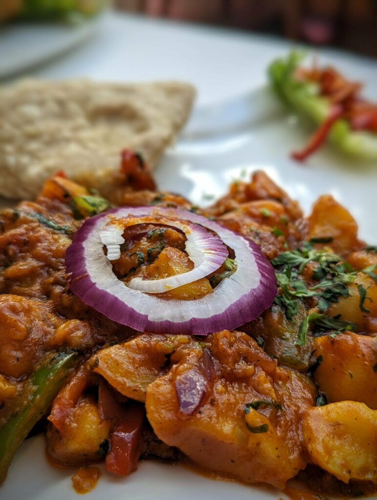 A vibrant vegetable curry topped with a raw red onion ring and fresh herbs, served on a white plate with flatbread.