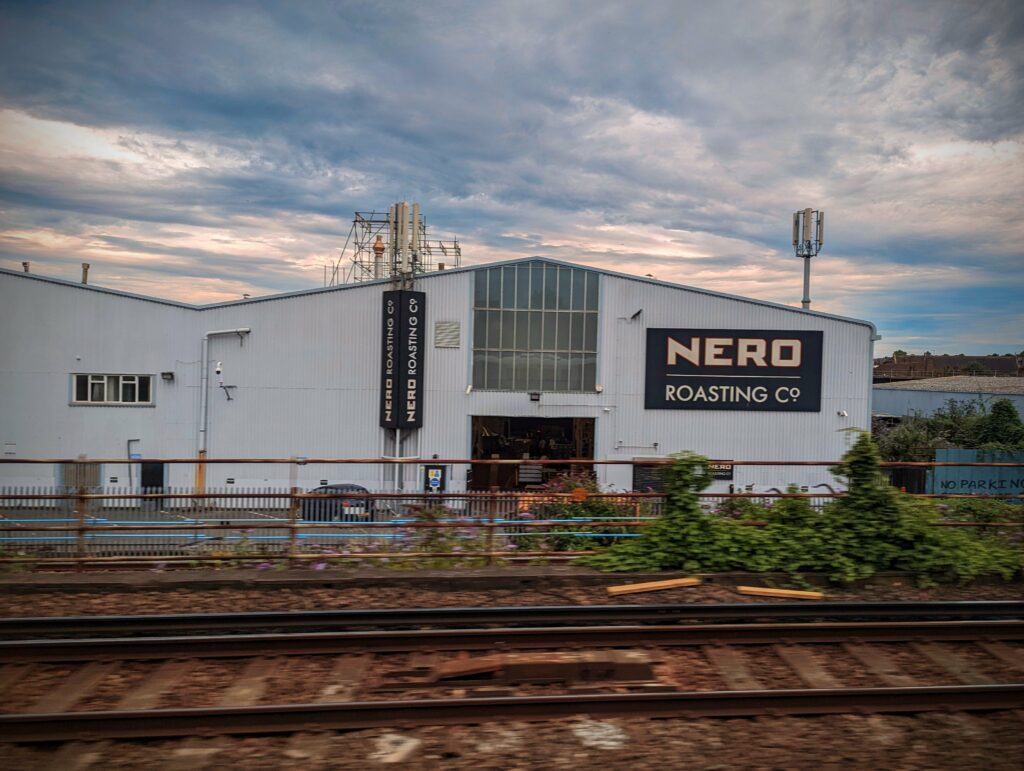 Nero Roasting Co industrial warehouse with large signage, seen from a passing train across railway tracks under a dramatic cloudy sky.