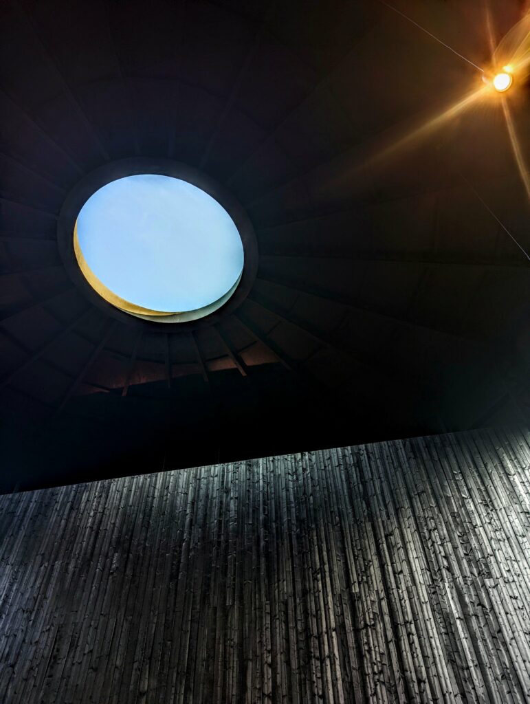 A circular oculus revealing a pale blue sky in a dark ribbed ceiling above a textured vertical wood wall with a bright light flare.