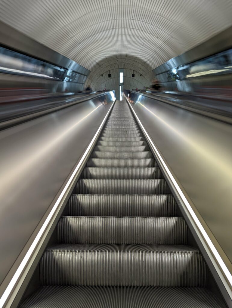 Symmetrical view looking up a long escalator in a modern subway tunnel with a curved ribbed ceiling and motion-blurred metal walls.