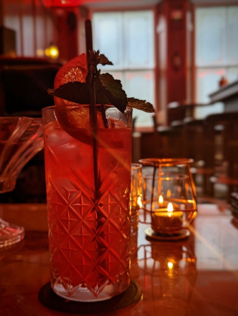Red cocktail in a cut-glass tumbler garnished with an orange slice and mint, set on a bar table beside a glowing tea light candle.