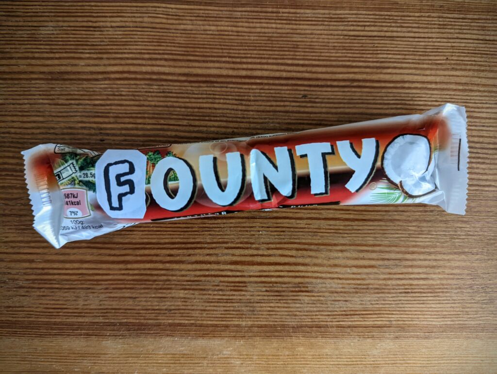 A Bounty chocolate bar modified with hand-drawn paper letters taped over the logo to read 'FOUNTY' on a wooden surface.