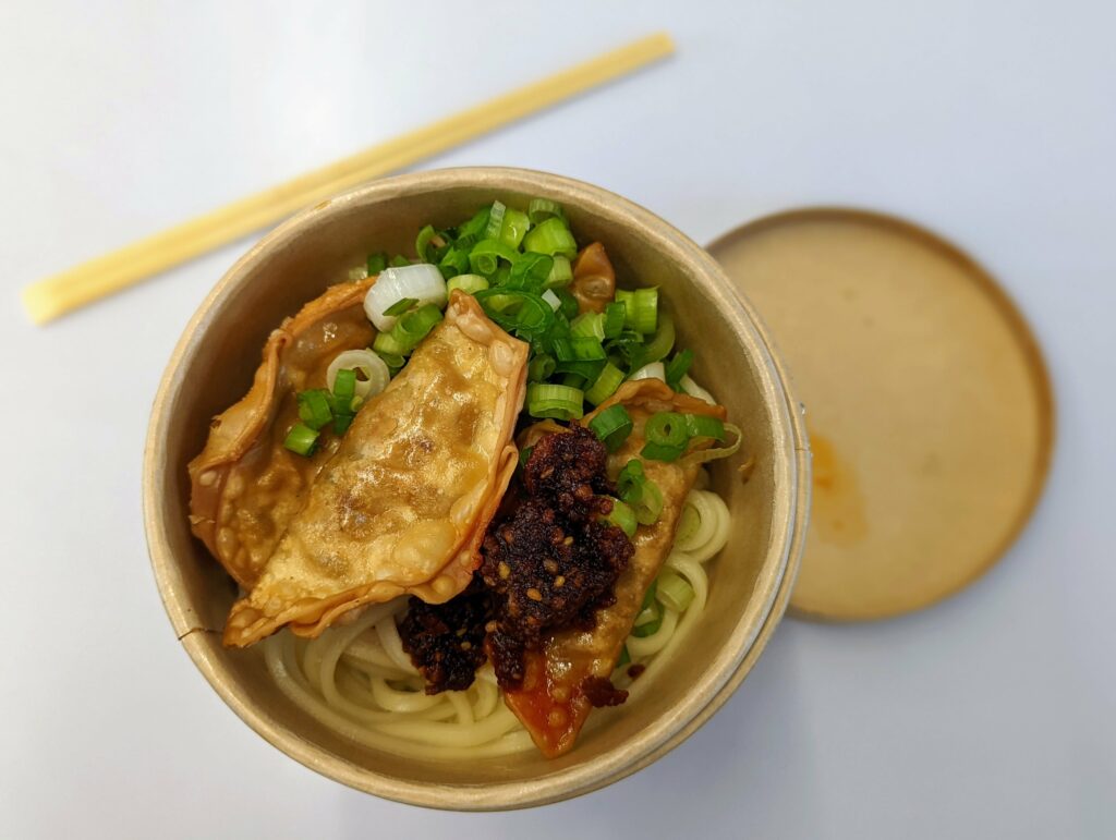 Fried dumplings and noodles topped with scallions and dark chili paste in a cardboard takeaway bowl next to wooden chopsticks.