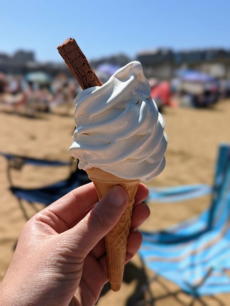 Vanilla soft-serve ice cream with a chocolate flake held against a sunny, blurred beach background with blue striped deckchairs.
