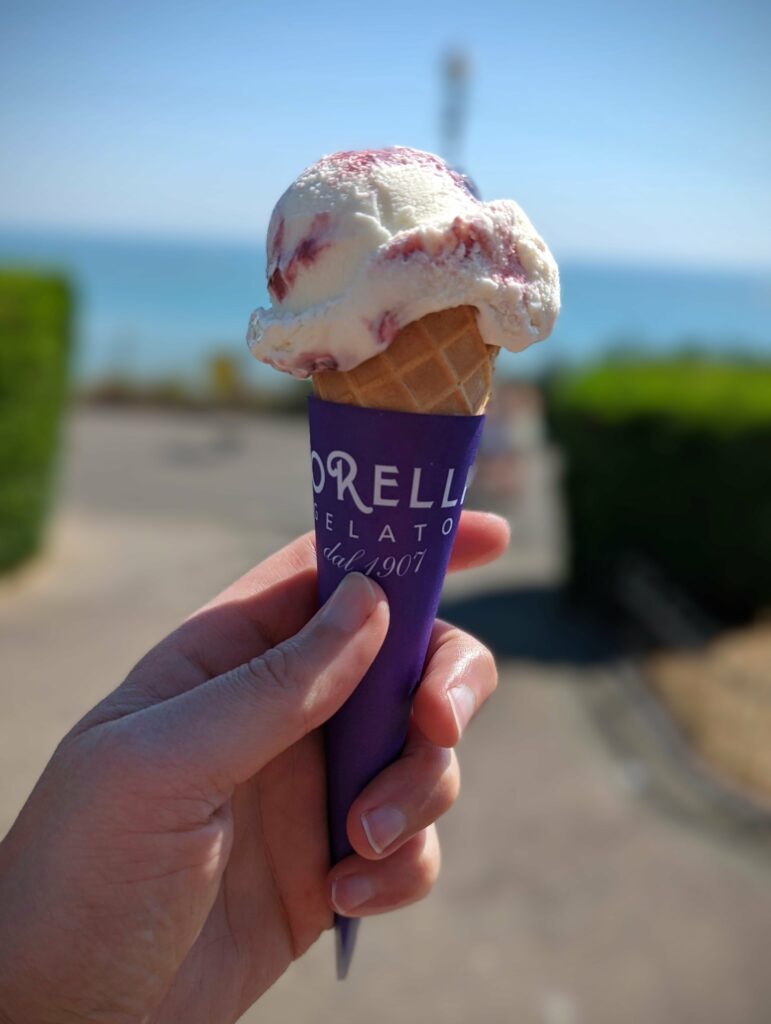 A hand holding a Morelli's gelato cone with a scoop of cherry ripple ice cream against a blurred sunny coastal background.