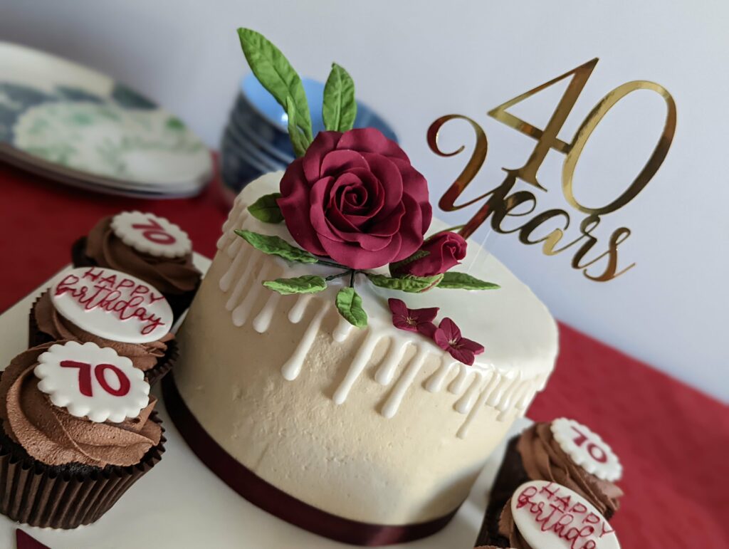 White drip cake topped with a red sugar rose and gold 40 Years topper, flanked by chocolate cupcakes with 70 and Happy Birthday decorations.