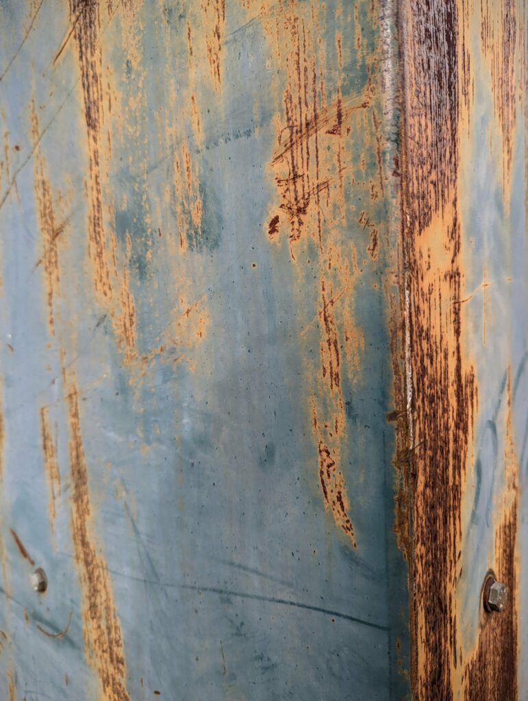 Vertical streaks of orange rust and deep scratches on a faded teal metal panel with silver bolts.