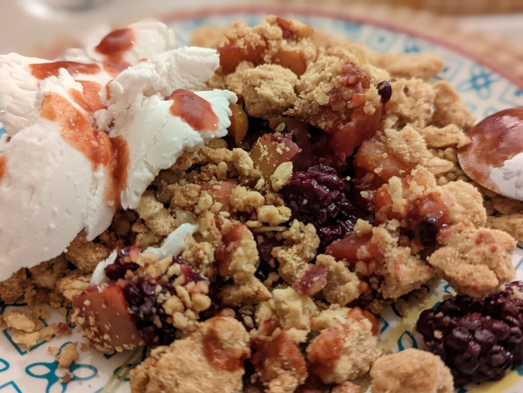 A blackberry and apple crumble topped with a dollop of cream and red fruit syrup on a blue patterned ceramic plate.