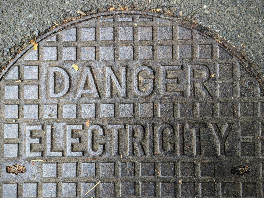 A cast iron utility cover in asphalt with the embossed warning DANGER ELECTRICITY on a raised square grid pattern.