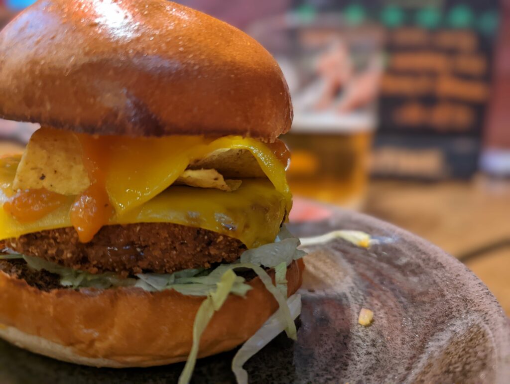 A burger with a breaded patty, melted cheese, tortilla chips, and lettuce in a brioche bun on a ceramic plate beside a blurred glass of beer.