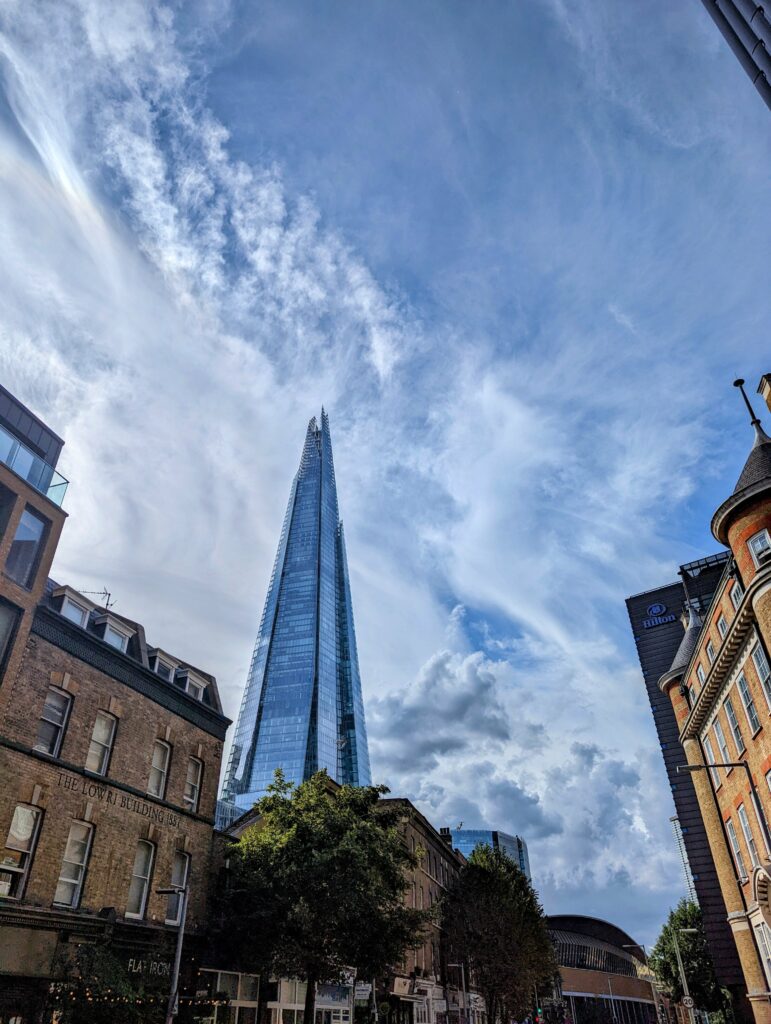 The glass Shard skyscraper towers over traditional brick buildings in London under a blue sky with dramatic wispy clouds.