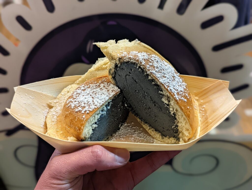 A hand holding a wooden boat tray with a sliced pancake filled with dense black sesame paste and dusted with powdered sugar.