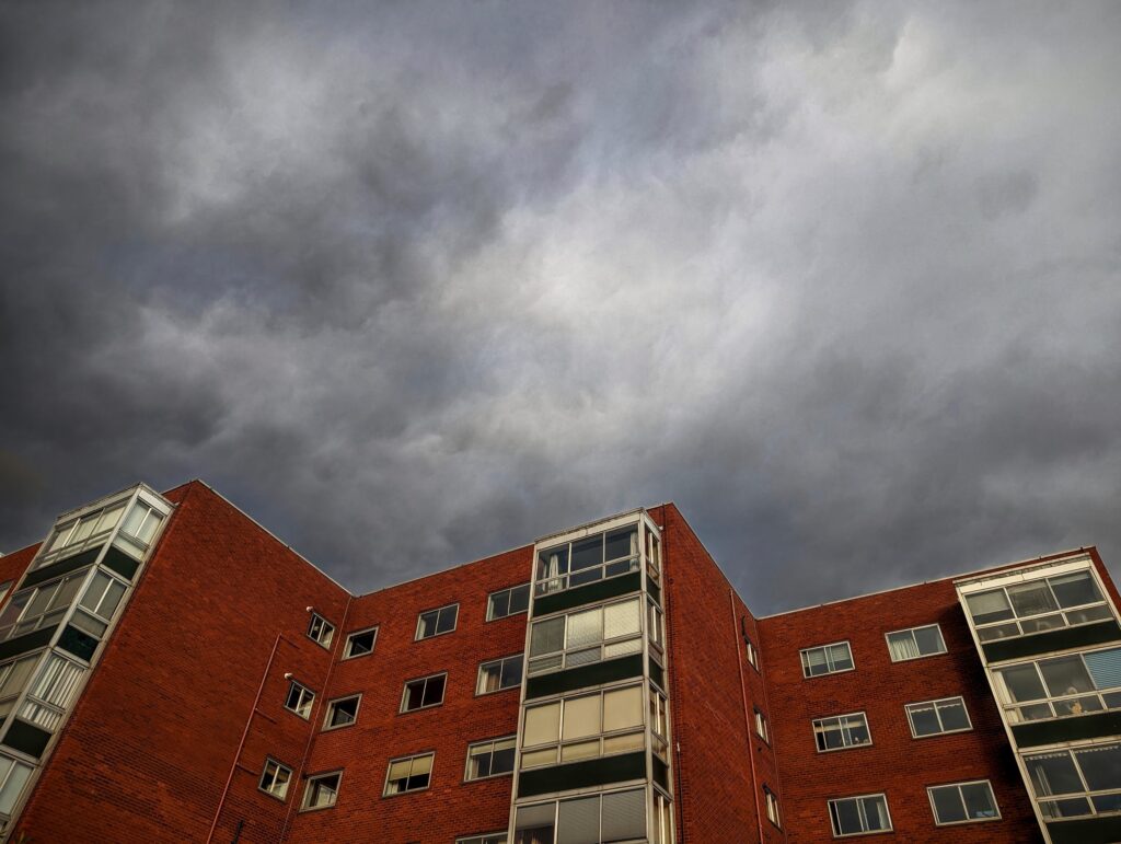 A red brick apartment building with glass-fronted balconies seen from a low angle against a dramatic, dark stormy sky.