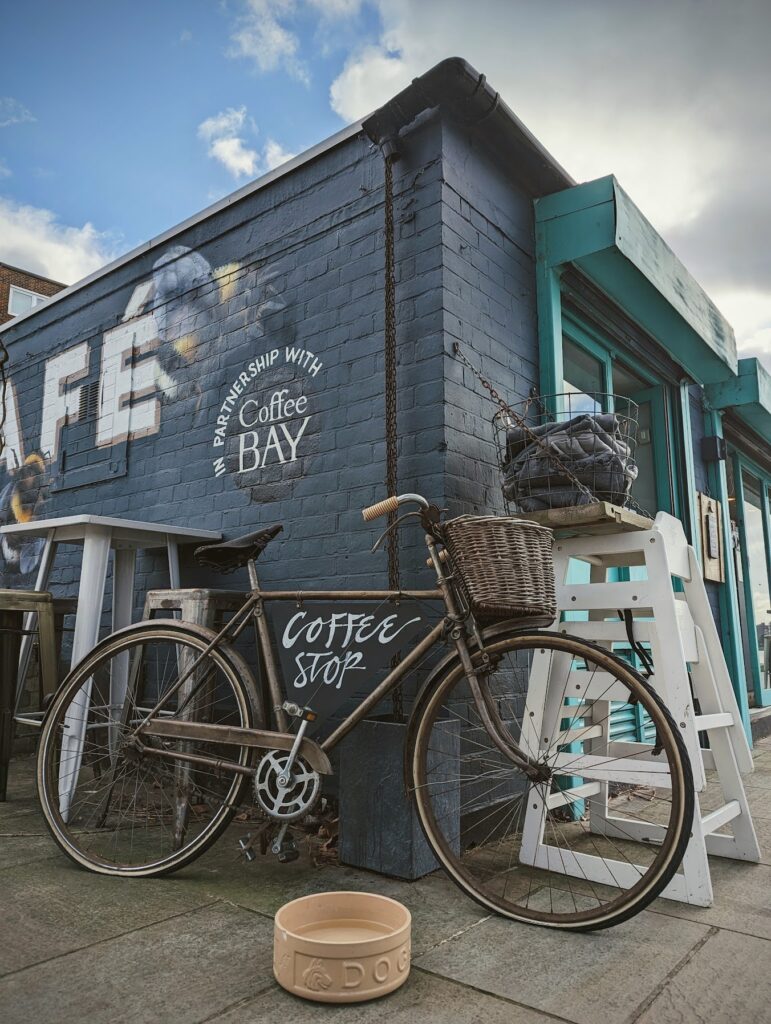 A vintage bicycle with a wicker basket parked against a blue brick cafe wall painted with bee murals and a Coffee BAY sign.