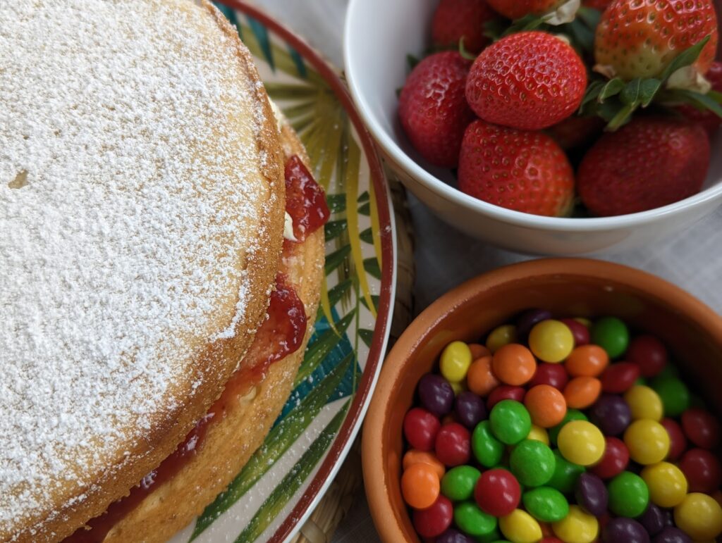 A Victoria sponge cake dusted with powdered sugar beside small bowls of fresh strawberries and colorful candy.