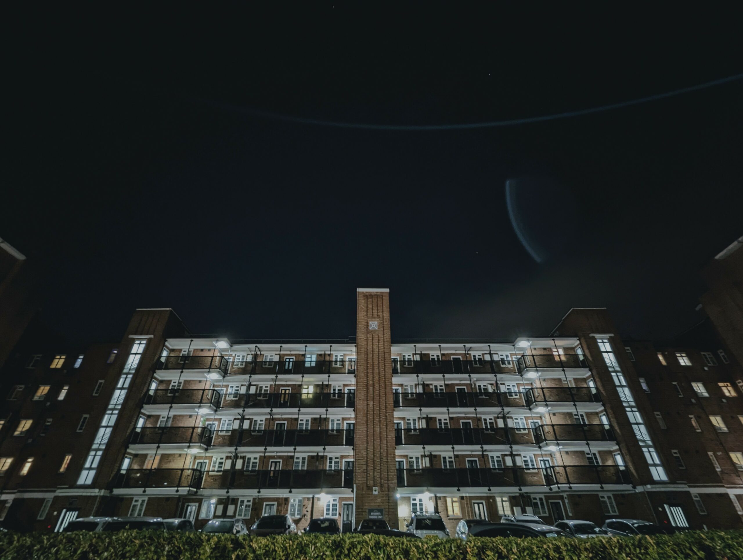 Symmetrical low-angle view of a brick apartment block at night with illuminated balconies and windows against a dark sky.
