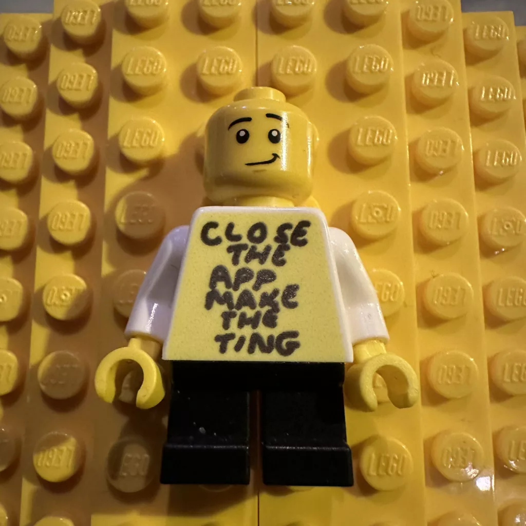 A smirk-faced LEGO minifigure on a yellow baseplate with handwritten text on its torso reading: CLOSE THE APP MAKE THE TING.