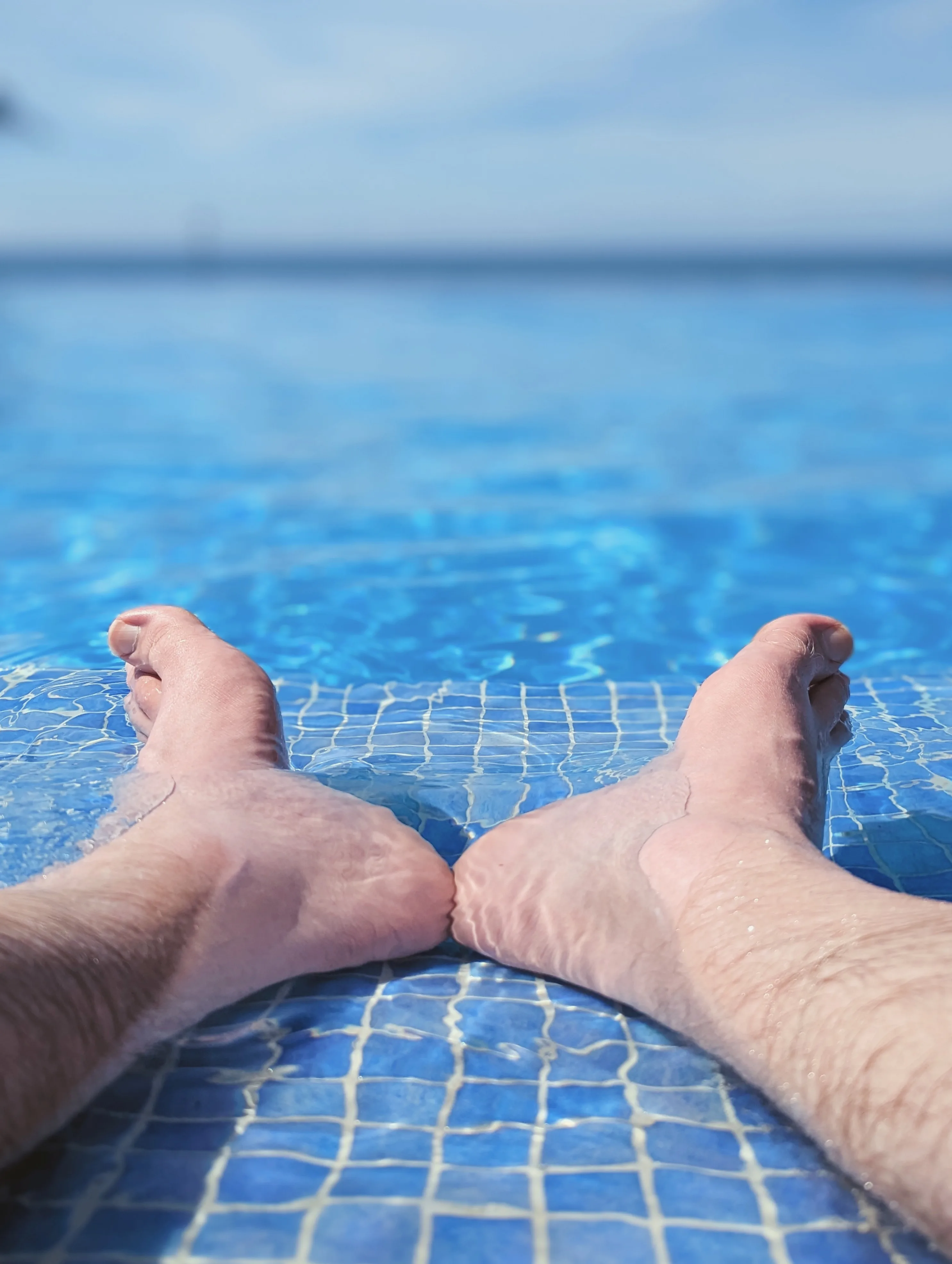 Two feet resting in the shallow water of a blue tiled swimming pool, looking out toward a blurred horizon line and clear sky.