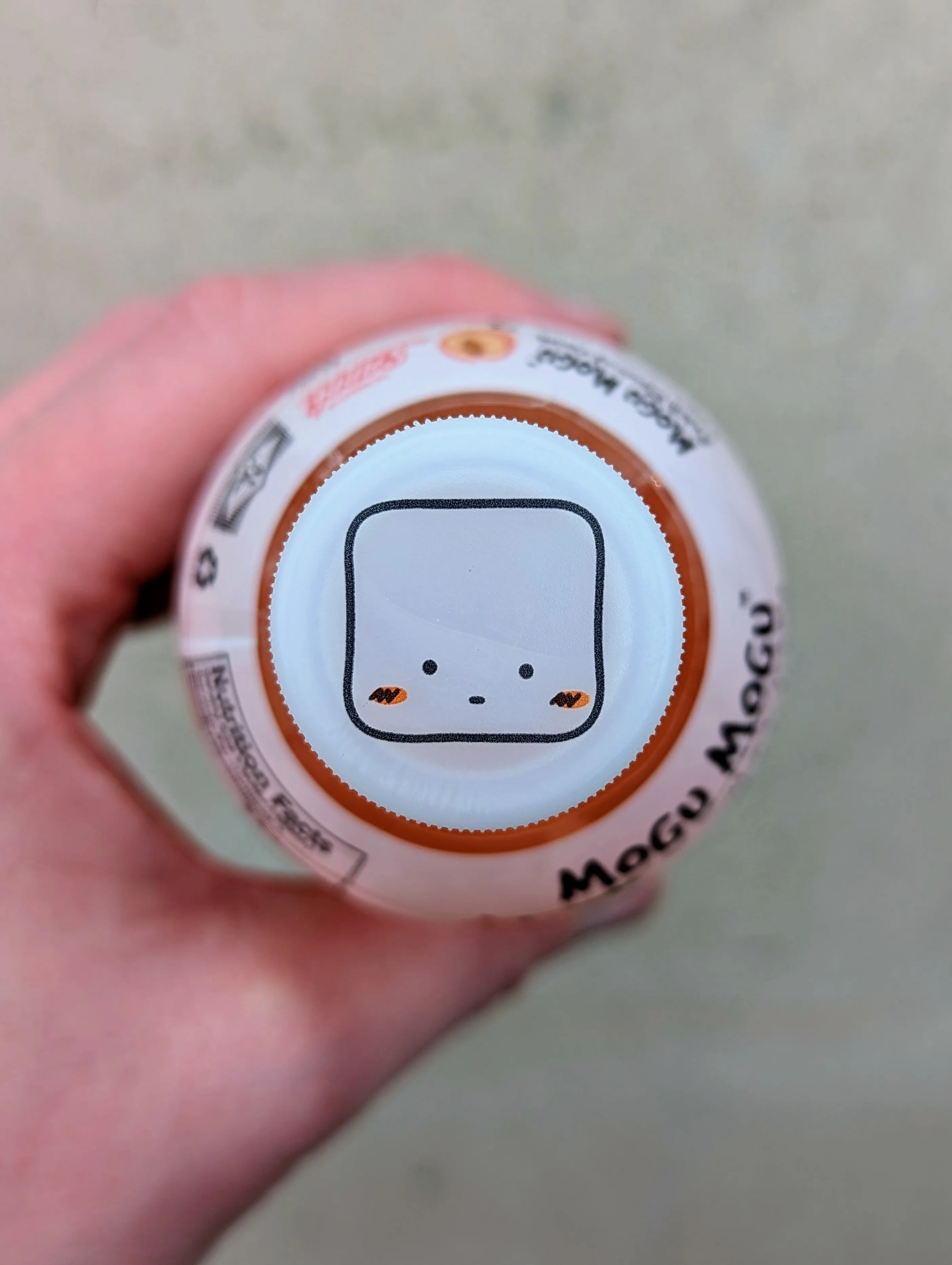 A minimalist square character with dot eyes and orange cheeks printed on the white cap of a Mogu Mogu drink bottle held in a hand.