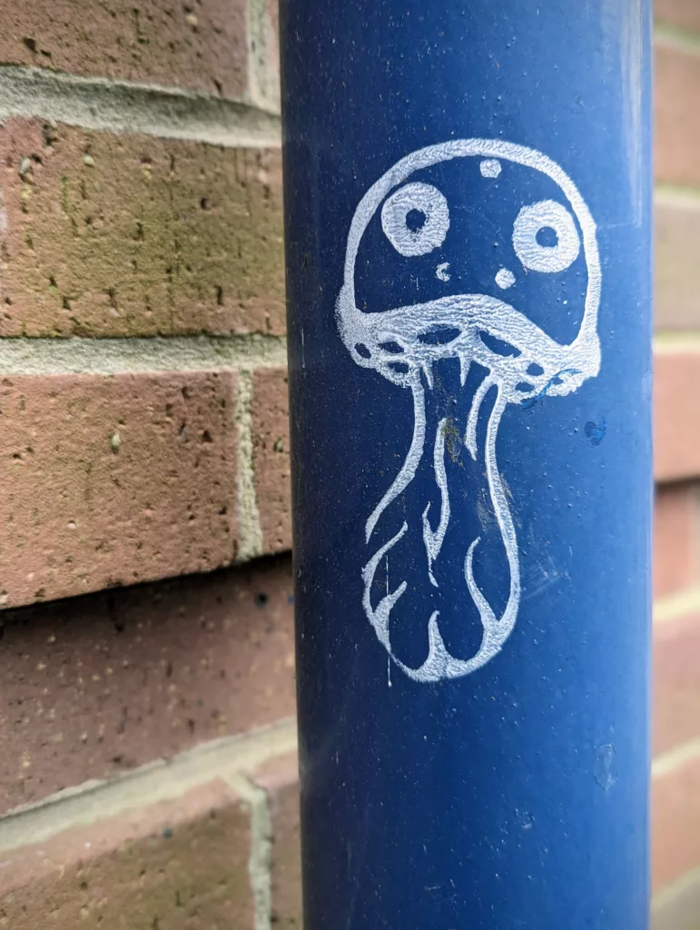 A white line drawing of a cartoon mushroom with large eyes on a blue downpipe beside a brick wall.