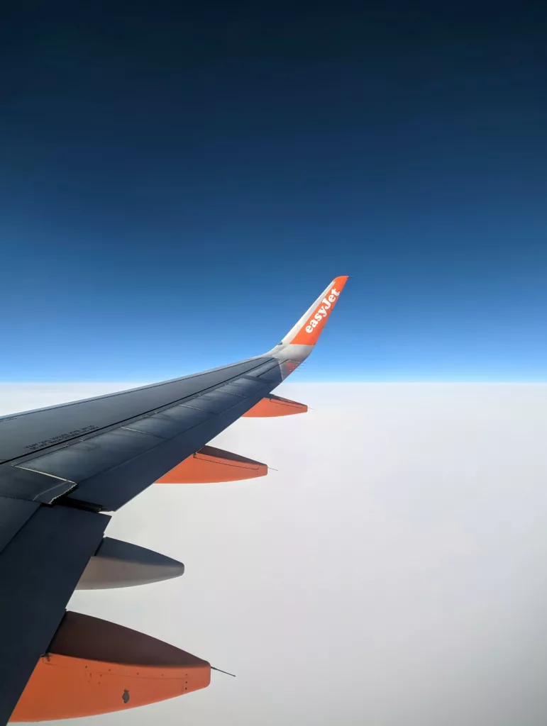 The wing of an easyJet airplane with its orange wingtip flying above a blanket of white clouds under a deep blue sky.