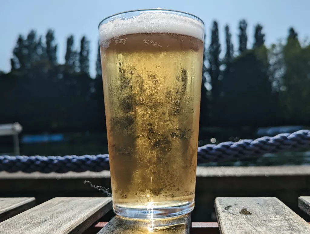 A sunlit glass of golden beer with a white head on a wooden table, against a blurred background of tall trees and a thick blue rope.