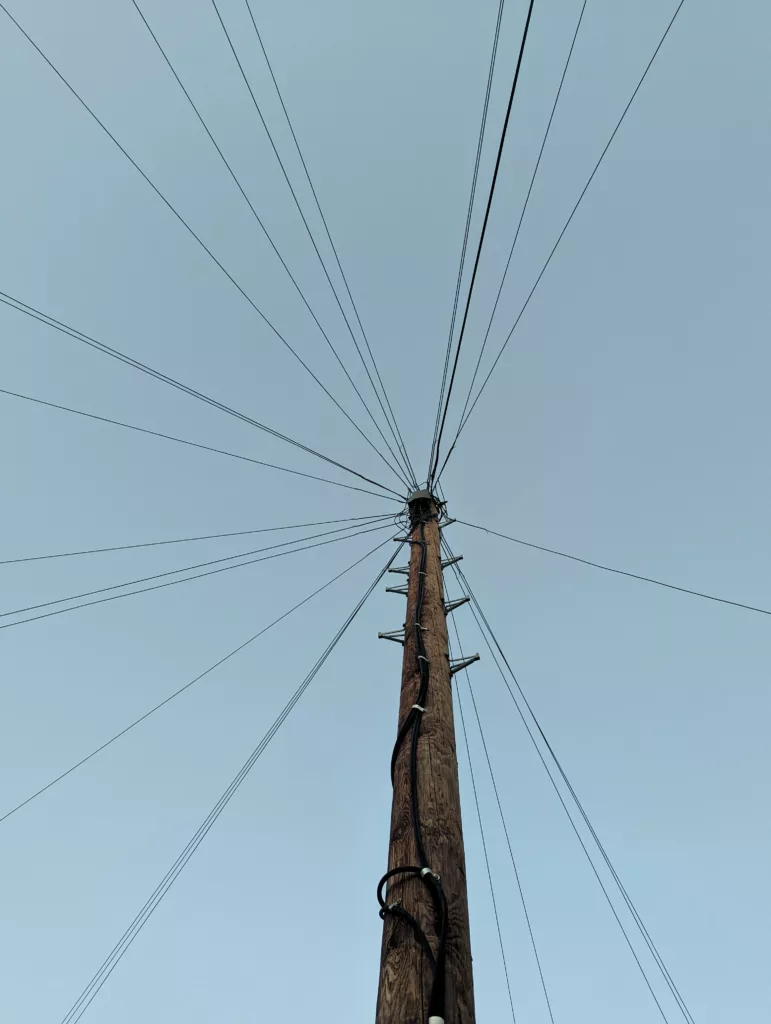 A wooden utility pole viewed from below, with numerous cables radiating outwards like spokes against a pale blue sky.