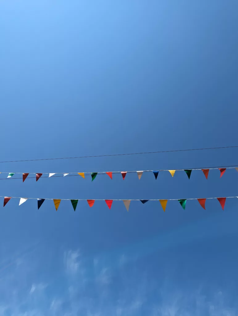 Two rows of colorful triangular bunting flags stretched across a clear, bright blue sky with wispy clouds at the bottom.