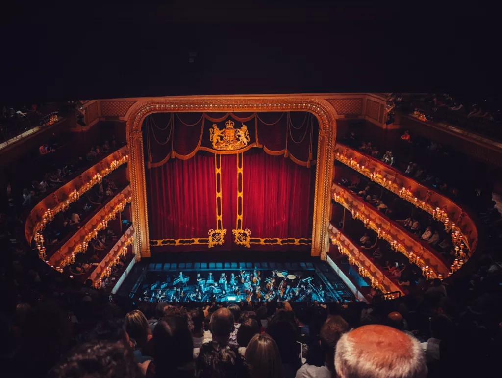 Ornate theatre interior with red stage curtains and a royal crest, seen from the balcony overlooking an orchestra pit and lit tiers of seating.