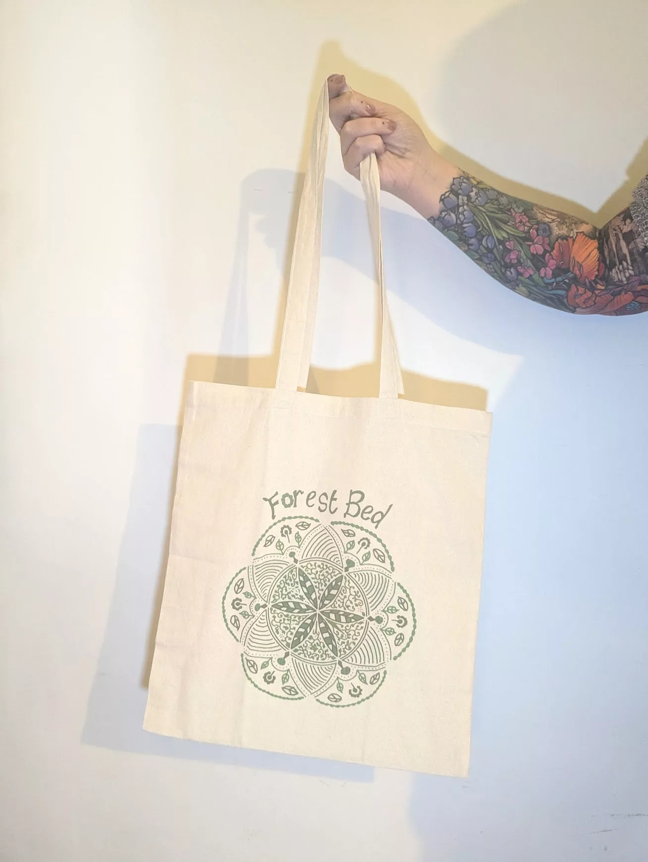 A tattooed hand holding a cream tote bag featuring a green circular mandala design and the text Forest Bed.