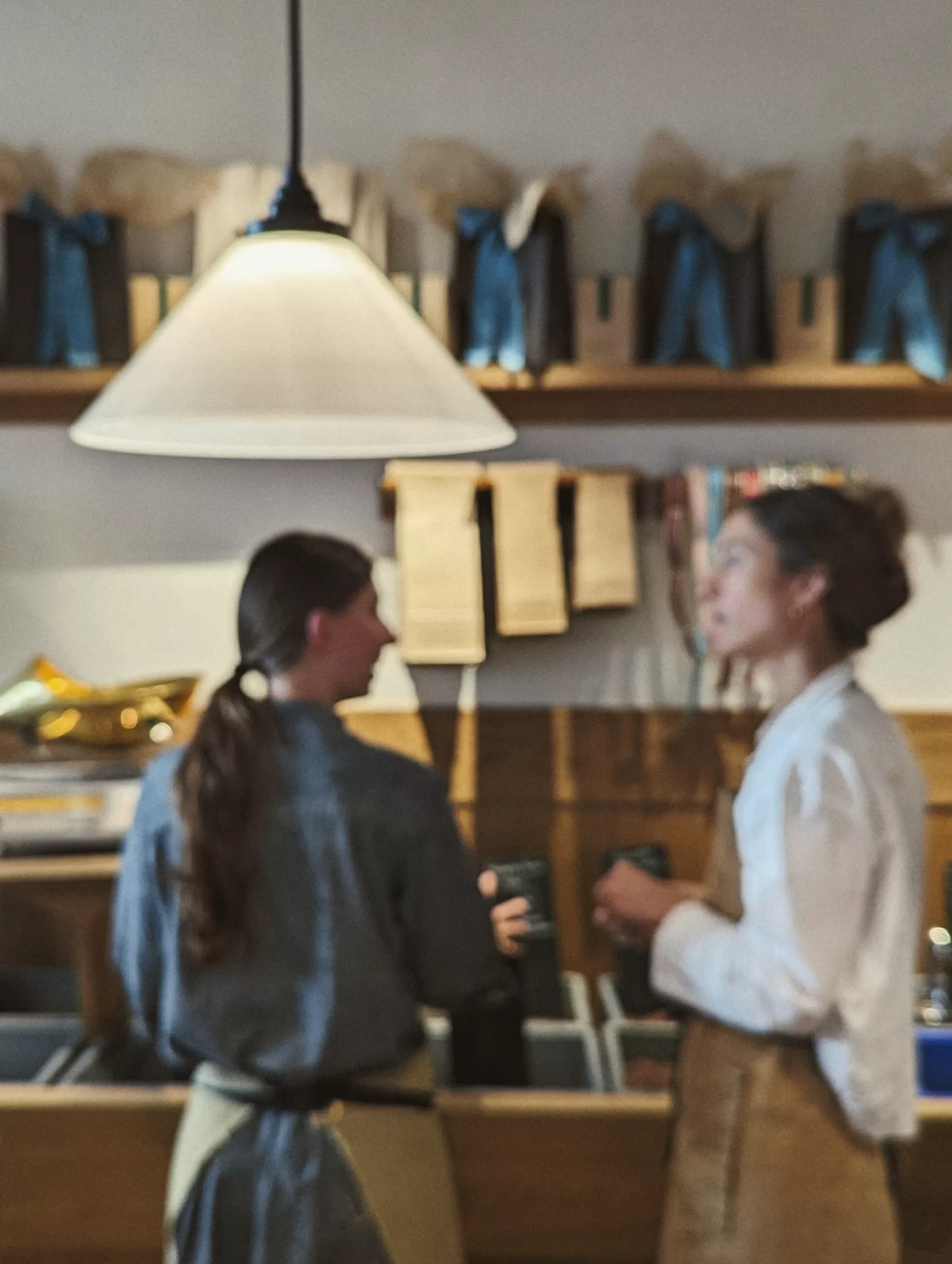 Two women in aprons talking behind a shop counter, blurred in the background of a prominent white conical pendant lamp.