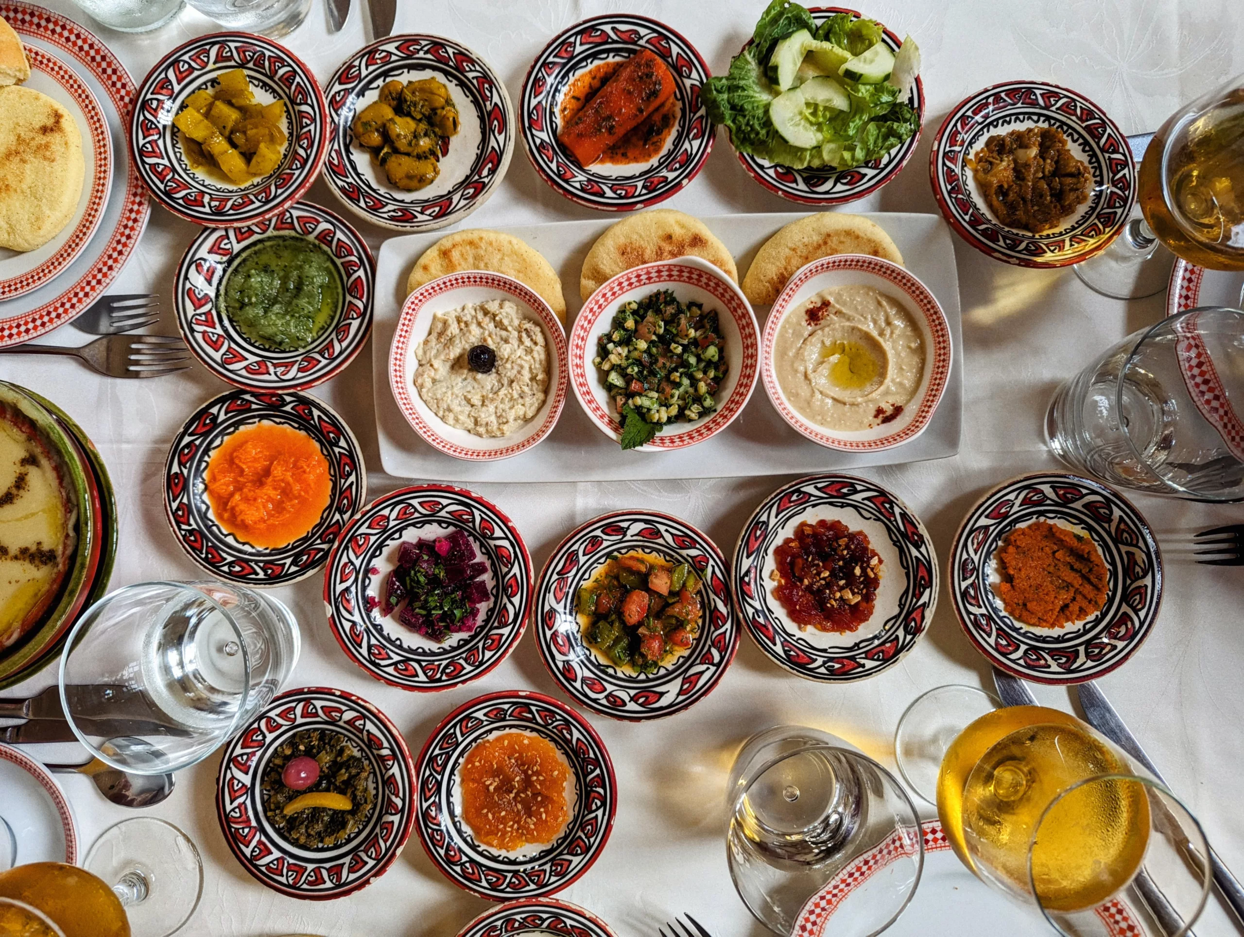 Assorted Mediterranean meze dishes, dips, and salads in patterned ceramic bowls on a white tablecloth with flatbread and wine glasses.