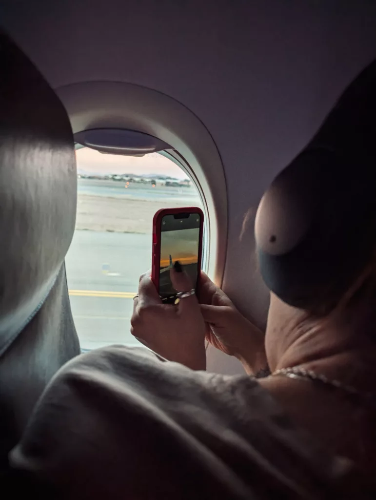 A passenger wearing headphones captures a sunset over an airport runway through an airplane window using a smartphone.