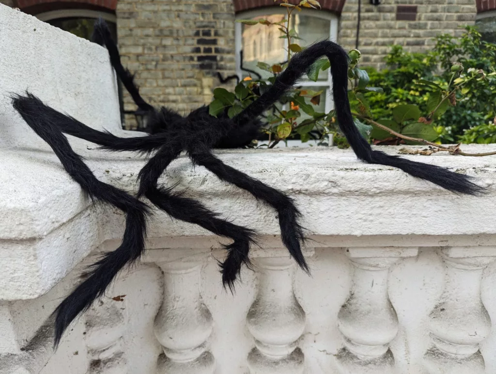 Large black fuzzy spider decoration with long legs draped over a white stone balustrade against a brick building.