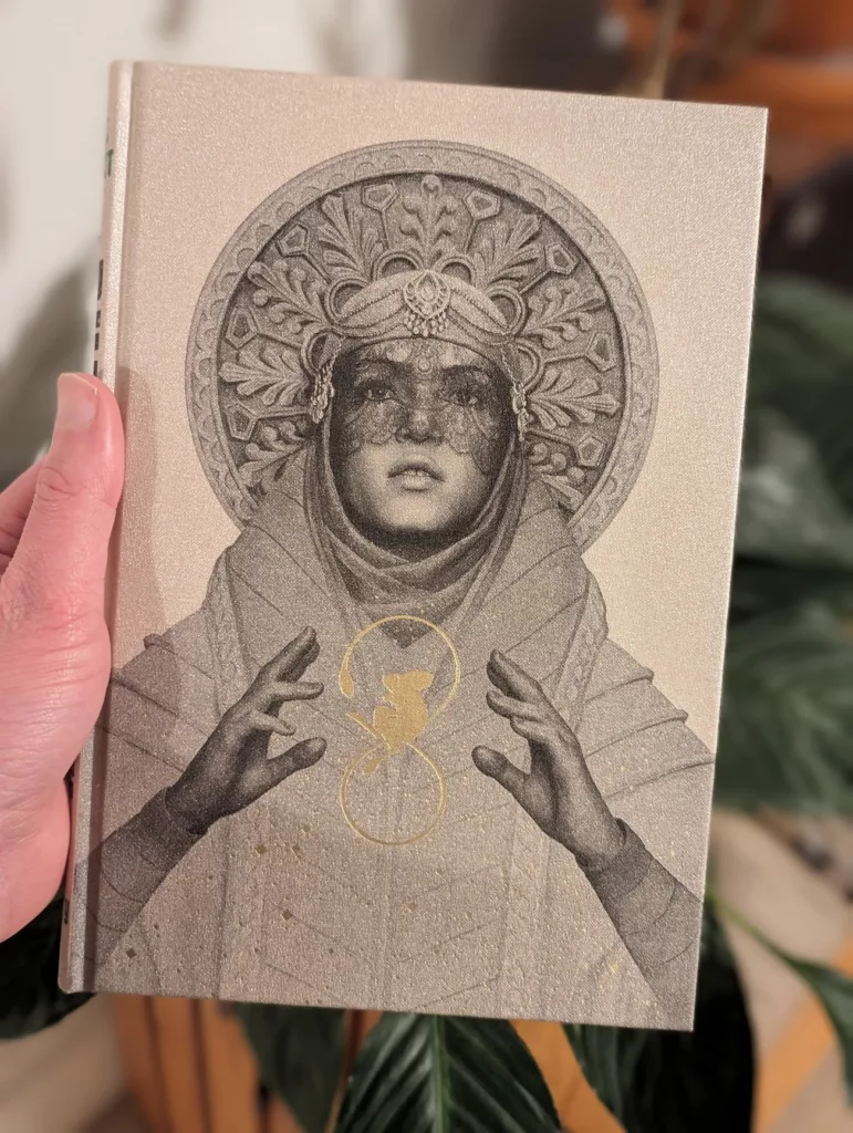 A hand holds a book with a shimmering cover featuring an ornate illustration of a woman in a regal headdress with a gold sigil between her hands.