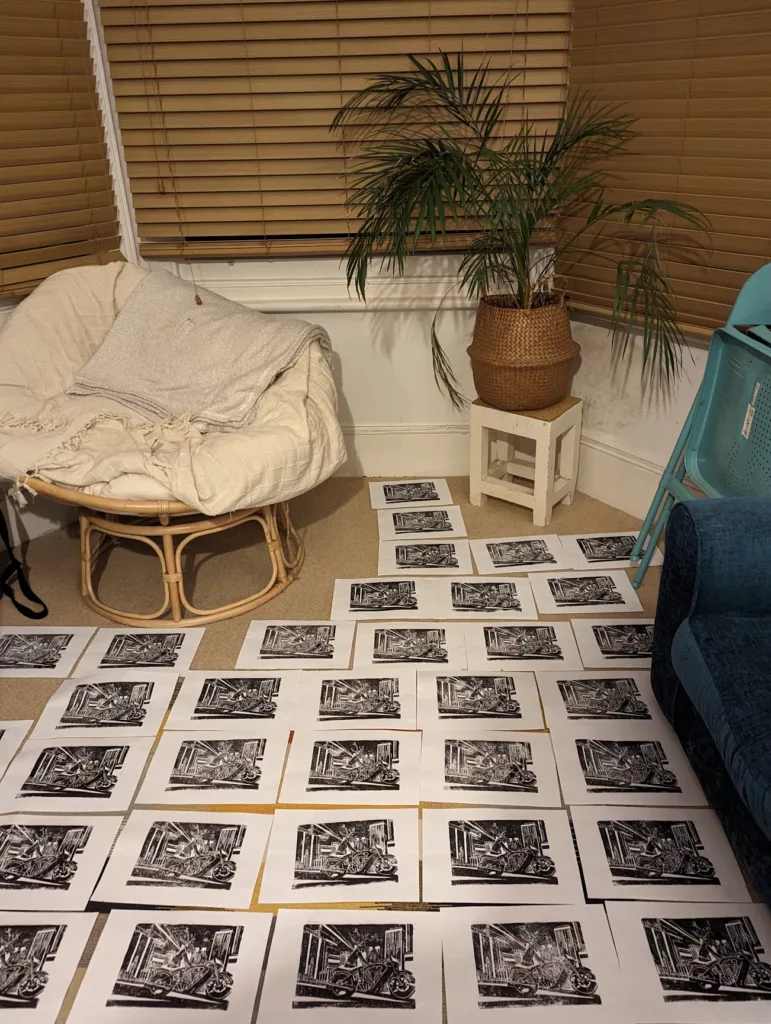 Numerous black and white linocut prints of a motorcycle drying on a carpeted floor between a papasan chair and a potted palm tree.