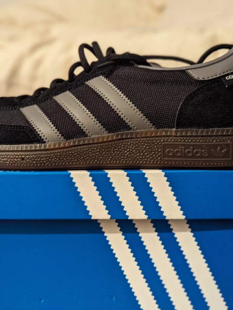 A black Adidas sneaker with three grey stripes and a gum sole resting on its blue and white striped shoebox.