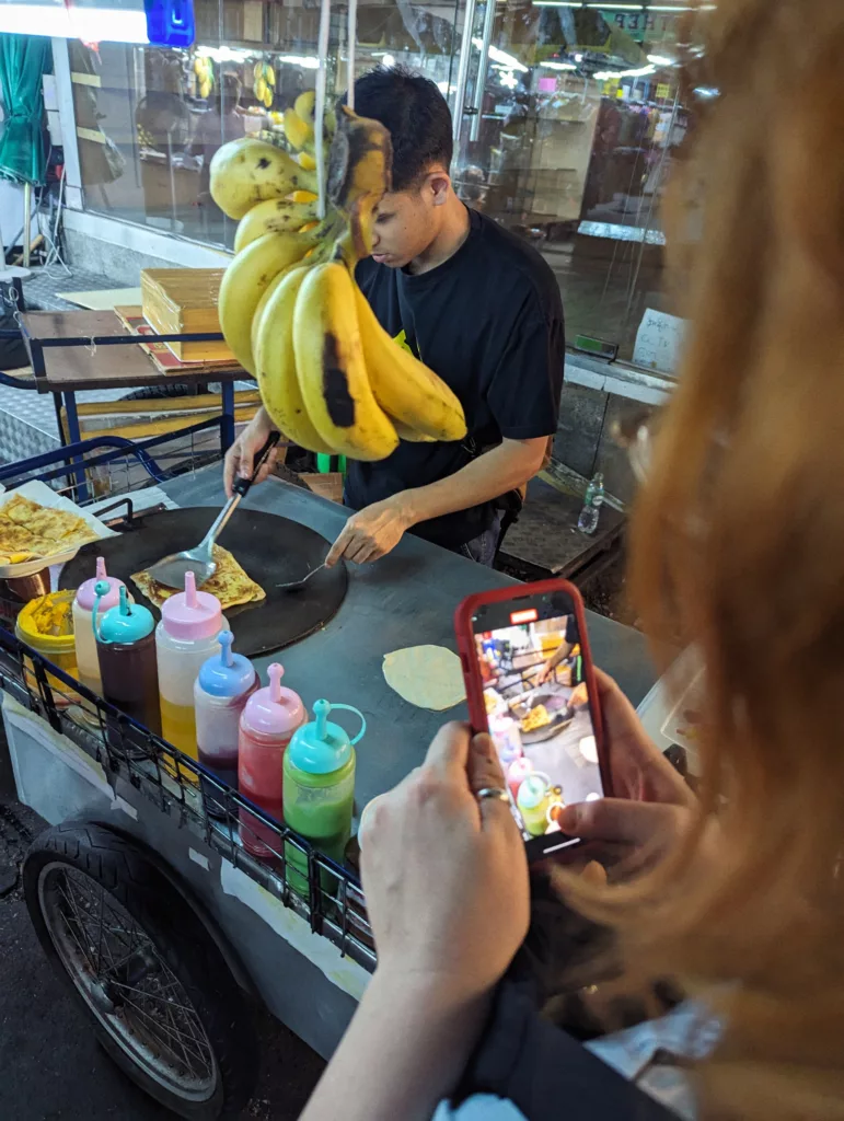 A street vendor cooking on a griddle behind colorful sauce bottles and hanging bananas while a person films the action on a smartphone.