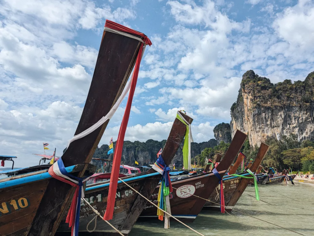 Traditional Thai longtail boats decorated with colorful ribbons moored along a beach with limestone cliffs and a cloudy sky.