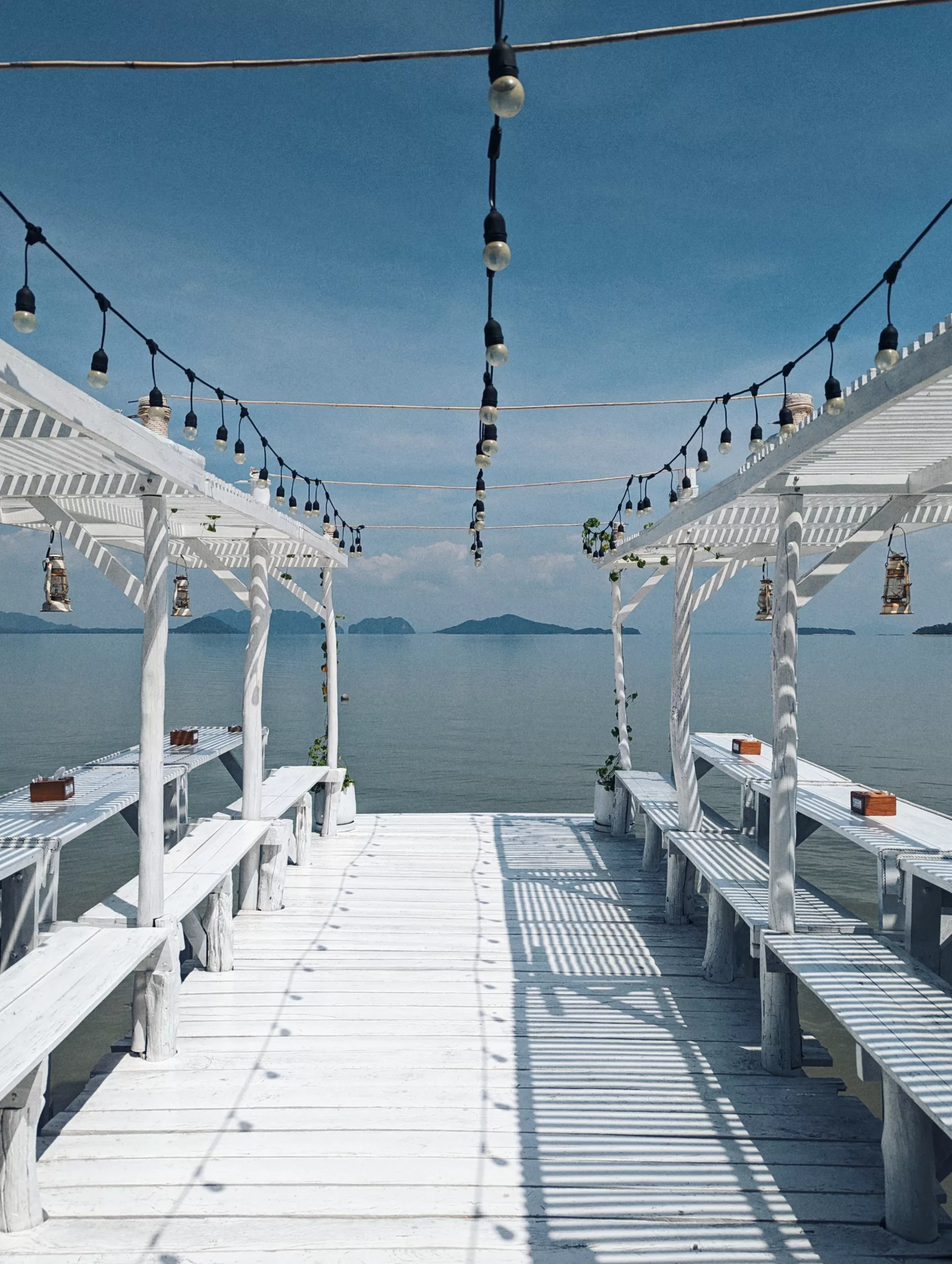 White wooden pier with outdoor seating under pergolas and festoon lights overlooking a calm sea and distant islands under a clear blue sky.