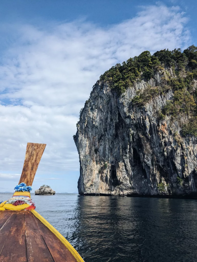 Decorated prow of a longtail boat heading towards a towering limestone cliff rising from the sea under a blue sky.