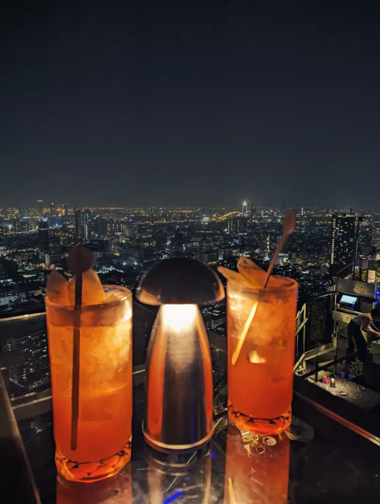 Two orange cocktails and a small table lamp on a rooftop bar overlooking a vast, illuminated city skyline at night.
